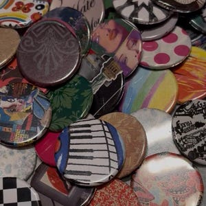 May include: A collection of assorted round buttons with various designs. The buttons feature images such as a piano keyboard, floral patterns, and abstract art. Colors include black, white, red, pink, and green. Some buttons have text.