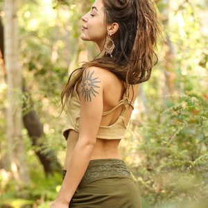 Peut inclure: Une femme aux longs cheveux bruns et un tatouage de soleil sur le bras porte un haut court marron et une jupe verte avec une bordure en dentelle. Elle se tient dans une forêt avec une végétation verte.