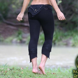 Puede incluir: Una mujer lleva un top corto verde con capucha y leggings negros con un cinturón negro con detalles dorados. Los leggings tienen encaje en la parte inferior. Está de pie en una zona cubierta de hierba con un cuerpo de agua en el fondo.