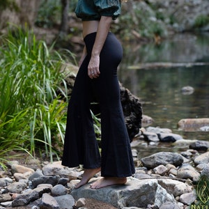 May include: A woman wearing a green off-the-shoulder top and black flared pants stands in a forest setting. She is standing on rocks near a stream.
