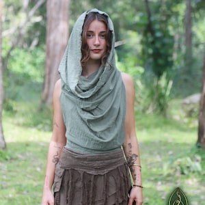 Può includere: Un top senza maniche color verde salvia con cappuccio, scollo drappeggiato e bordi grezzi, abbinato a una minigonna marrone scuro con strisce di tessuto sovrapposte e orlo sfrangiato. Il logo Nature Spirit è visibile nell'angolo in basso a destra.