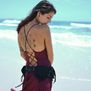 Op de afbeelding: Een vrouw draagt een bordeauxrode jurk met een zwarte leren riem met zakken en franjes. De jurk heeft een gesnorte rug en de vrouw heeft een tatoeage op haar rug. Ze staat op een strand met de zee op de achtergrond.