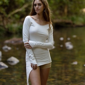 Puede incluir: Una camisa blanca de manga larga con cuello redondo y un dobladillo ligeramente asimétrico. La camisa está hecha de una tela suave y elástica y tiene un ajuste holgado. La modelo lleva la camisa con unos pantalones cortos beige y está de pie en un entorno boscoso.