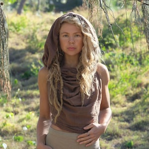 May include: A woman with long blonde hair wearing a brown hooded top with a gathered neckline. She is standing in a grassy field with trees in the background.