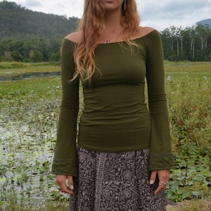 Può includere: Un top verde oliva con spalle scoperte e maniche lunghe, abbinato a una gonna lunga e fluida. La gonna presenta un motivo floreale marrone scuro. L'outfit è indossato all'aperto, con un lago e montagne sullo sfondo.
