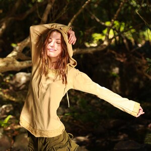 Könnte beinhalten: Eine Frau trägt ein hellbraunes Kapuzenpullover mit langen Ärmeln und einen grünen Rock. Sie steht in einem Wald mit grünen Bäumen und Sonnenlicht, das durch die Blätter scheint.