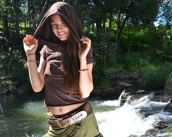 May include: A woman wearing a brown hooded top, a green and brown skirt with a brown belt, and brown trousers. She is standing in front of a waterfall.
