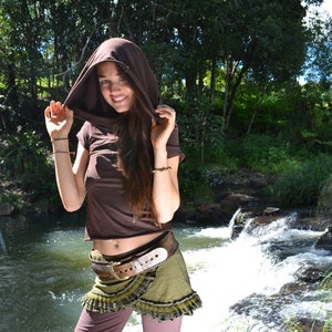 Op de afbeelding: Een vrouw draagt een bruin getailleerd vest met capuchon, een groen en bruin rok met een bruine riem en een bruine broek. Ze staat voor een waterval.