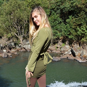 Può includere: Una donna indossa un top avvolgente verde oliva a maniche lunghe con leggings marroni. È in piedi su una roccia vicino a una cascata.