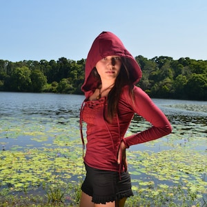 Könnte beinhalten: Eine Frau trägt ein rotes Kapuzenoberteil mit Schnürung vorne und schwarze Shorts. Sie steht vor einem See mit Seerosen.