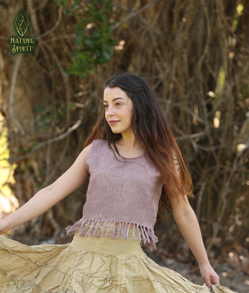 May include: A woman wearing a mauve sleeveless top with fringe trim and a beige tiered skirt. The top is made of a textured fabric. The woman is standing in a natural setting with trees and foliage in the background.