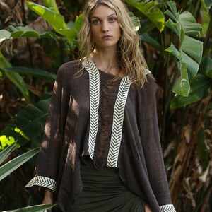 Pu&ograve; includere: Un poncho marrone con bordature bianche a chevron, indossato da una persona con lunghi capelli biondi. Il poncho ha un bordo sfrangiato ed &egrave; realizzato in un tessuto leggero. La persona &egrave; in piedi in un lussureggiante giardino verde.