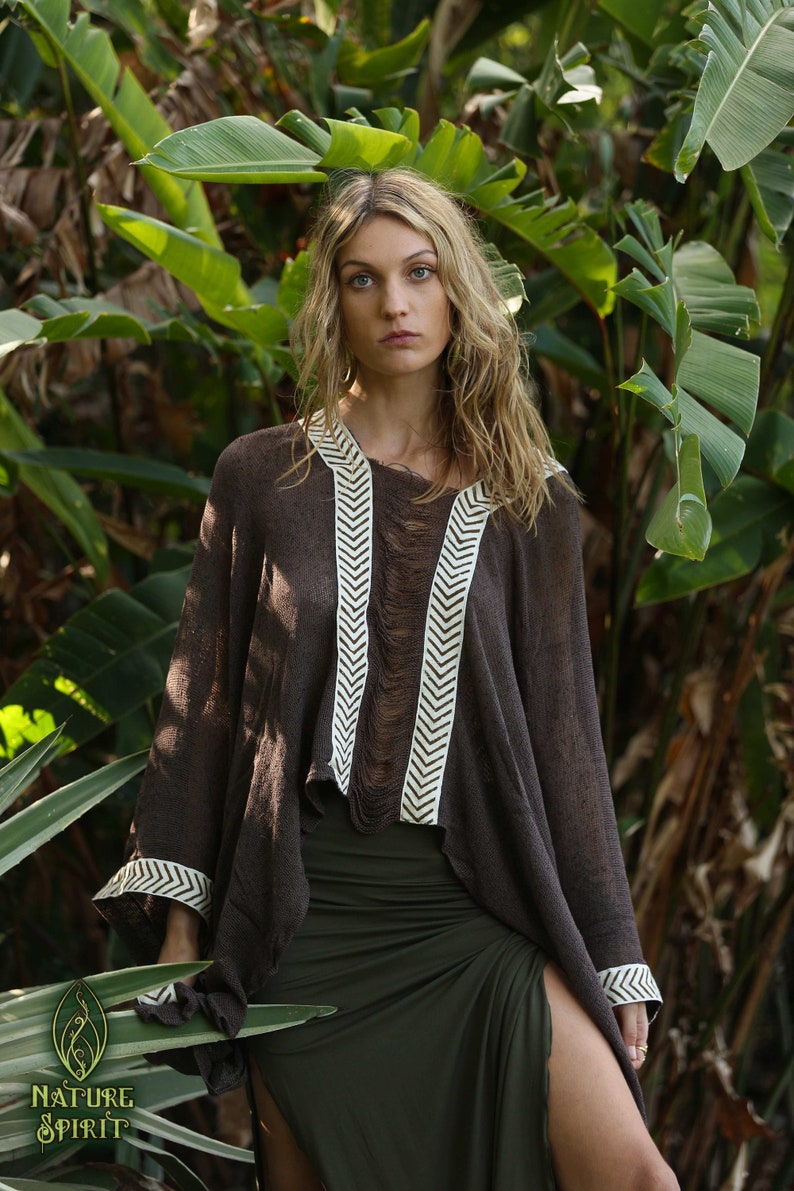 Pu&ograve; includere: Un poncho marrone con bordature bianche a chevron, indossato da una persona con lunghi capelli biondi. Il poncho ha un bordo sfrangiato ed &egrave; realizzato in un tessuto leggero. La persona &egrave; in piedi in un lussureggiante giardino verde.
