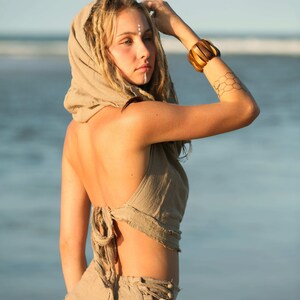 Op de afbeelding: Een vrouw draagt een lichtbruin getailleerd vest met een kap en een bijpassende rok met een rafelige zoom. Ze staat op een strand met de zee op de achtergrond. Het vest is aan de achterkant vastgebonden en de rok heeft een vetersluiting. Ze draagt een houten armband om haar pols.