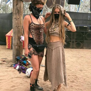 Op de afbeelding: Twee mensen dragen gezichtsmaskers en festivalkleding. Een persoon draagt een luipaardprint top met een zwarte riem en zwarte laarzen. De andere persoon draagt een bruine capuchontrui met een bruine rok en is op blote voeten.