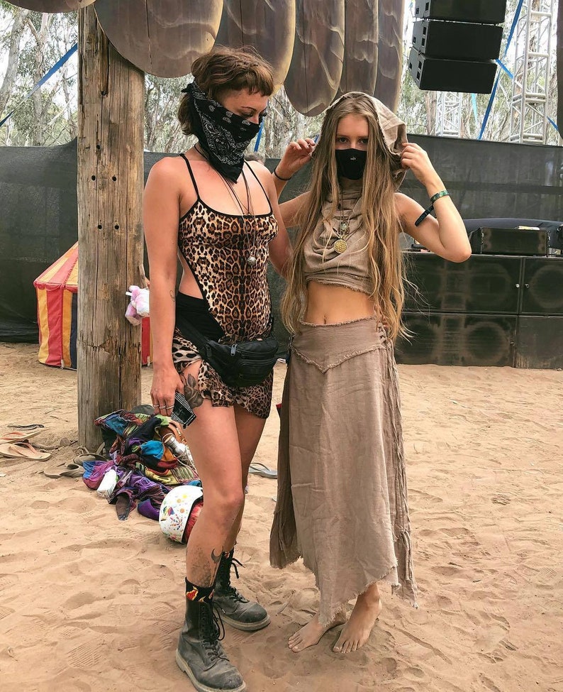Op de afbeelding: Twee mensen dragen gezichtsmaskers en festivalkleding. Een persoon draagt een luipaardprint top met een zwarte riem en zwarte laarzen. De andere persoon draagt een bruine capuchontrui met een bruine rok en is op blote voeten.