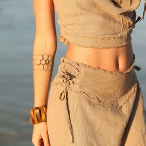 Op de afbeelding: Een vrouw draagt een lichtbruin linnen top en rok met een honingraat-tatoeage op haar arm. De rok heeft een vetersluiting aan de zijkant.