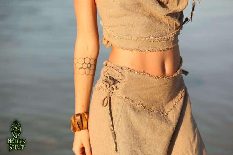 Op de afbeelding: Een vrouw draagt een lichtbruin linnen top en rok met een honingraat-tatoeage op haar arm. De rok heeft een vetersluiting aan de zijkant.