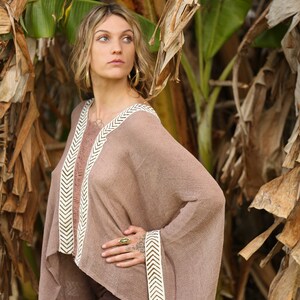 Pu&ograve; includere: Una donna indossa un poncho marrone con bordi a chevron bianchi e marroni. Il poncho &egrave; realizzato in un tessuto leggero e traspirante e ha una vestibilit&agrave; rilassata. La donna &egrave; in piedi in un ambiente da giardino con piante di banana sullo sfondo.