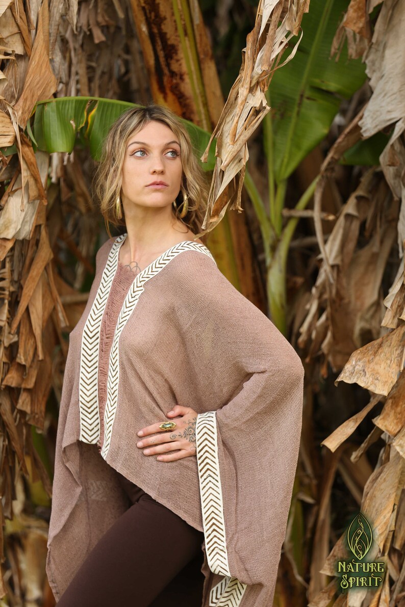 Pu&ograve; includere: Una donna indossa un poncho marrone con bordi a chevron bianchi e marroni. Il poncho &egrave; realizzato in un tessuto leggero e traspirante e ha una vestibilit&agrave; rilassata. La donna &egrave; in piedi in un ambiente da giardino con piante di banana sullo sfondo.