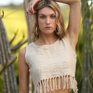 May include: A woman wearing a white crocheted crop top with fringe detailing. The top is sleeveless and has a round neckline. The woman has a tattoo on her stomach.