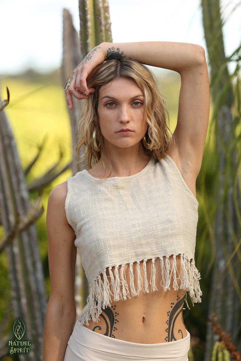 May include: A woman wearing a white crocheted crop top with fringe detailing. The top is sleeveless and has a round neckline. The woman has a tattoo on her stomach.