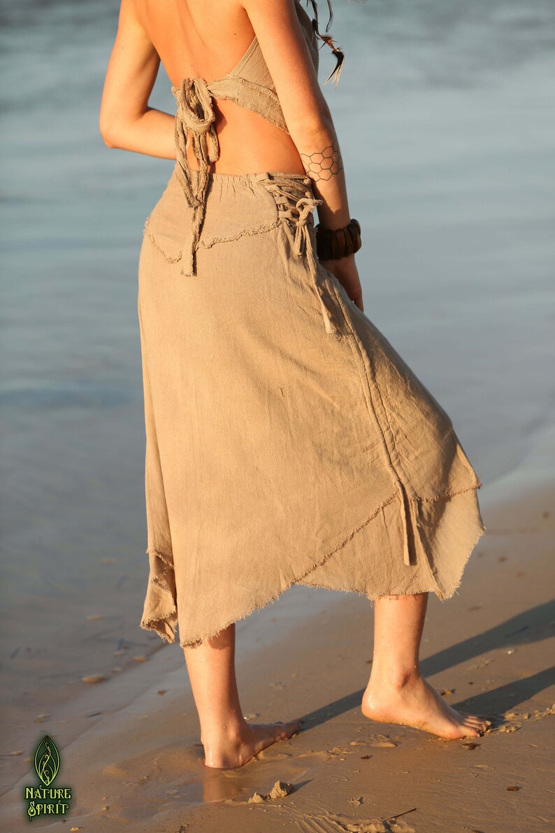 Op de afbeelding: Een vrouw draagt een lichtbruine linnen rok met een rauwe zoom en een strik in de taille. De rok is zwierig en heeft een losse pasvorm. De vrouw staat op een zandstrand.