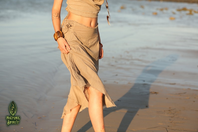 Op de afbeelding: Een vrouw draagt een lichtbruine, zwierige rok met een rafelige zoom. De rok is in de taille gebonden en heeft een losse, casual pasvorm. De vrouw staat op een zandstrand met de zee op de achtergrond.