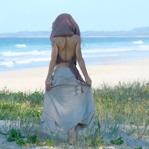 Op de afbeelding: Een vrouw draagt een lichtbruine rok en een lichtroze top met een capuchon. Ze staat op een zandstrand met groen gras en de zee op de achtergrond.