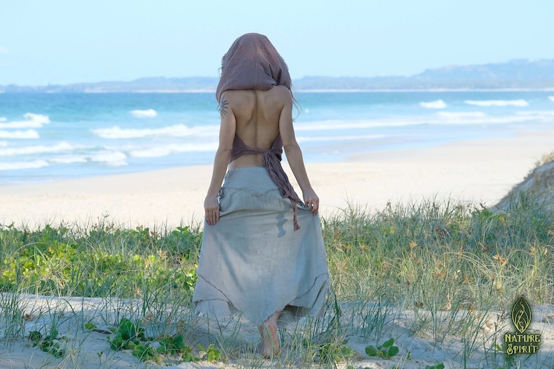 Op de afbeelding: Een vrouw draagt een lichtbruine rok en een lichtroze top met een capuchon. Ze staat op een zandstrand met groen gras en de zee op de achtergrond.