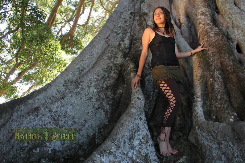 May include: A woman wearing a black lace top and brown pants with lace detailing stands in front of a large tree trunk. The tree trunk is covered in moss and has a rough texture. The woman is looking up at the sky. The text "Nature Spirit" is visible at the bottom of the image.