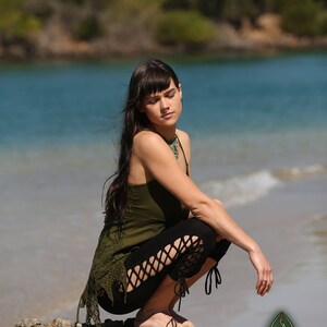 May include: A woman wearing a green tank top and black leggings with cutouts sits on a rock by the water. The leggings have a lace-up detail at the ankles. The woman has long dark hair and is wearing a necklace.