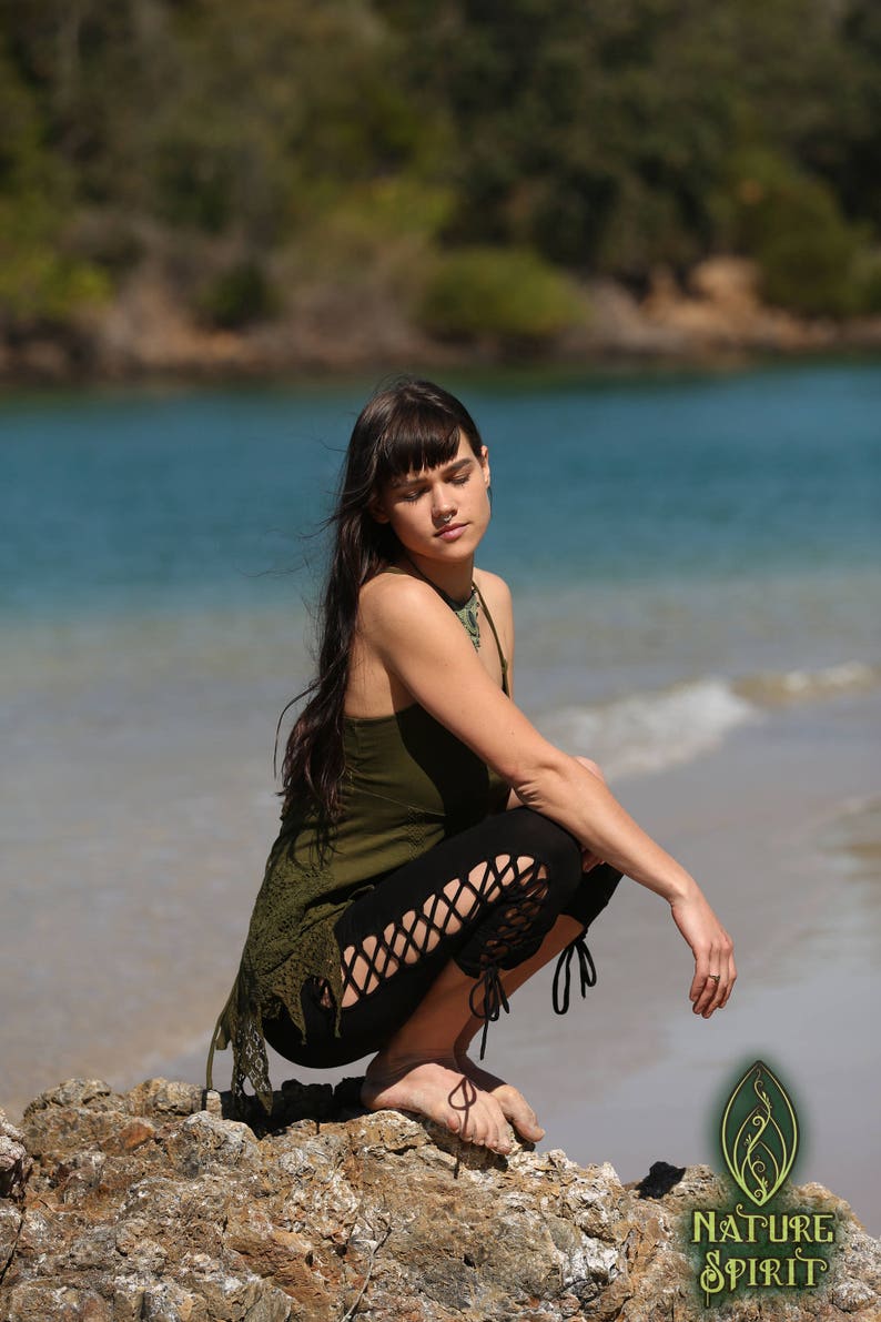 May include: A woman wearing a green tank top and black leggings with cutouts sits on a rock by the water. The leggings have a lace-up detail at the ankles. The woman has long dark hair and is wearing a necklace.