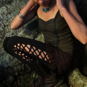 May include: A woman wearing a dark green top with a turquoise and silver necklace, black leggings with cutouts, and brown boots. She is sitting on a mossy rock.