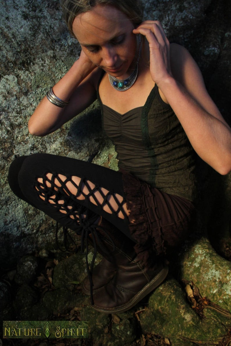 May include: A woman wearing a dark green top with a turquoise and silver necklace, black leggings with cutouts, and brown boots. She is sitting on a mossy rock.