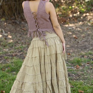May include: A woman wearing a mauve sleeveless top with a lace-up back and a brown tiered skirt with lace trim. The skirt is long and flowing, and the top is cropped. The woman is standing in a grassy area.