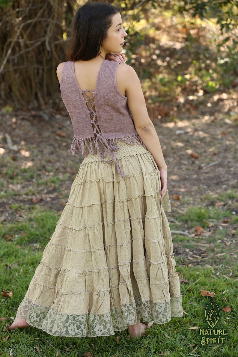 May include: A woman wearing a mauve sleeveless top with a lace-up back and a brown tiered skirt with lace trim. The skirt is long and flowing, and the top is cropped. The woman is standing in a grassy area.