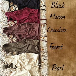 May include: Five tops in black, maroon, chocolate, forest green, and pearl. Each top has a crisscross design and is displayed on a white lace surface. The colours are labelled with handwritten text.