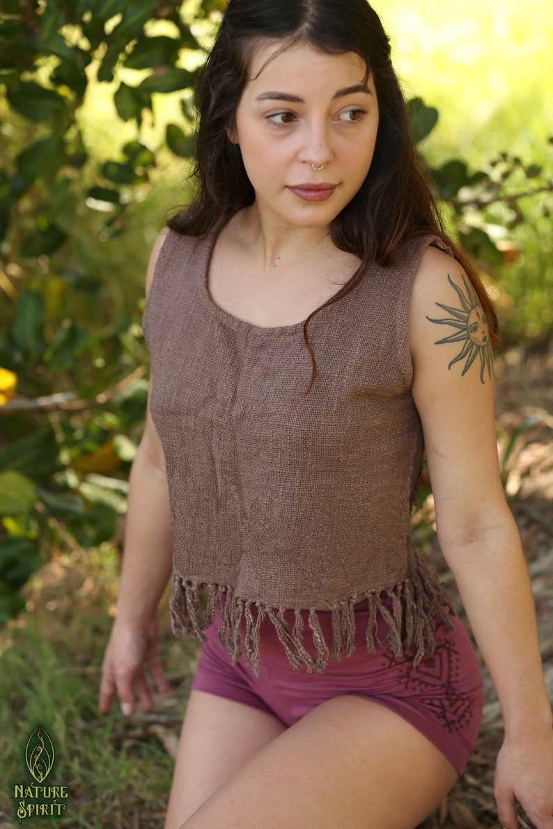 May include: A woman wearing a brown sleeveless top with fringe detailing and a pair of burgundy shorts with a geometric pattern. She has long brown hair and a nose ring.