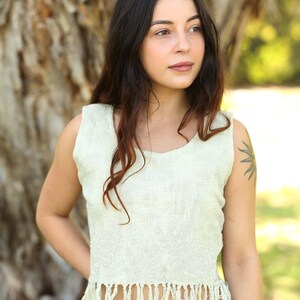 May include: A woman wearing a white sleeveless top with fringe trim. The top is made of a natural fiber and has a relaxed fit. The woman is standing in front of a tree.