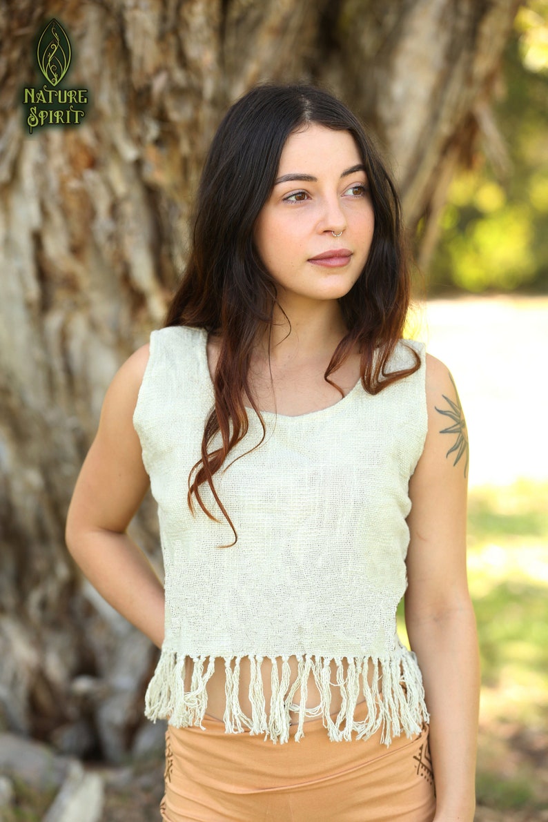 May include: A woman wearing a white sleeveless top with fringe trim. The top is made of a natural fiber and has a relaxed fit. The woman is standing in front of a tree.