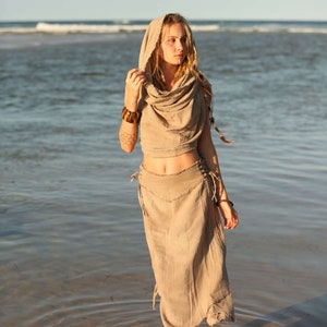 Op de afbeelding: Een vrouw die een lichtbruine linnen jurk draagt met een capuchon en een lange rok. Ze staat in ondiep water op een zandstrand.