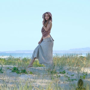 Op de afbeelding: Een vrouw in een lichtbruin gehooded top en een lange, vloeiende, lichtgrijze rok staat op een zandstrand met groen gras. De vrouw kijkt naar de camera. De tekst "NATURE SPIRIT" is zichtbaar in de rechterbenedenhoek van de afbeelding.