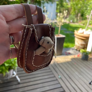 Op de afbeelding: Een klein bruin leren rugzakje met een gestikt ontwerp en een witte teddybeer die uit de zak kijkt.