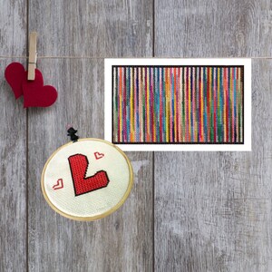 May include: A cross-stitch embroidery hoop with a red letter "L" and small red hearts. A framed artwork with vertical stripes of various colors hangs nearby. Two red heart cutouts are clipped to a string.