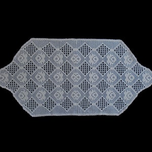 May include: White, handmade crochet table runner with a geometric pattern. The runner features a repeating design of squares and circles, creating an openwork texture. The edges are scalloped, adding a decorative touch. Ideal for adding a vintage or handmade aesthetic to a table.
