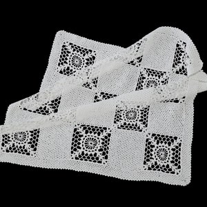 Può includere: Una tovaglietta bianca all'uncinetto con un motivo quadrato e disegni floreali centrali. La tovaglietta è piegata e drappeggiata, mettendo in mostra l'intricato traforo e la superficie strutturata su sfondo nero. L'articolo artigianale è fatto a mano.