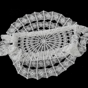 May include: A white, handmade lace doily with an intricate openwork design. The doily has a central oval shape surrounded by a circular pattern of geometric shapes and a scalloped edge. The delicate details are set against a black background.