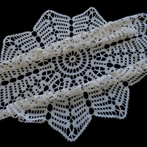 May include: A cream-coloured, handmade crochet doily with a starburst design. The intricate lace pattern features a central circular motif surrounded by geometric shapes and a scalloped edge. The doily is displayed against a black background.