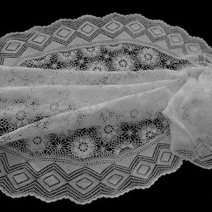 Op de afbeelding: Een witte kanten tafelloper met een ingewikkeld bloemenpatroon in het midden en een geometrische ruitrand langs de zijkanten. Het delicate textiel vertoont een complex opengewerkt ontwerp met geschulpte randen en fijn draadwerk.